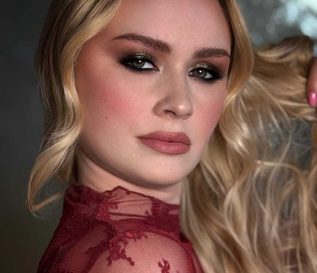 Young woman with wavy blonde hair, wearing dark eye makeup and red lace top.