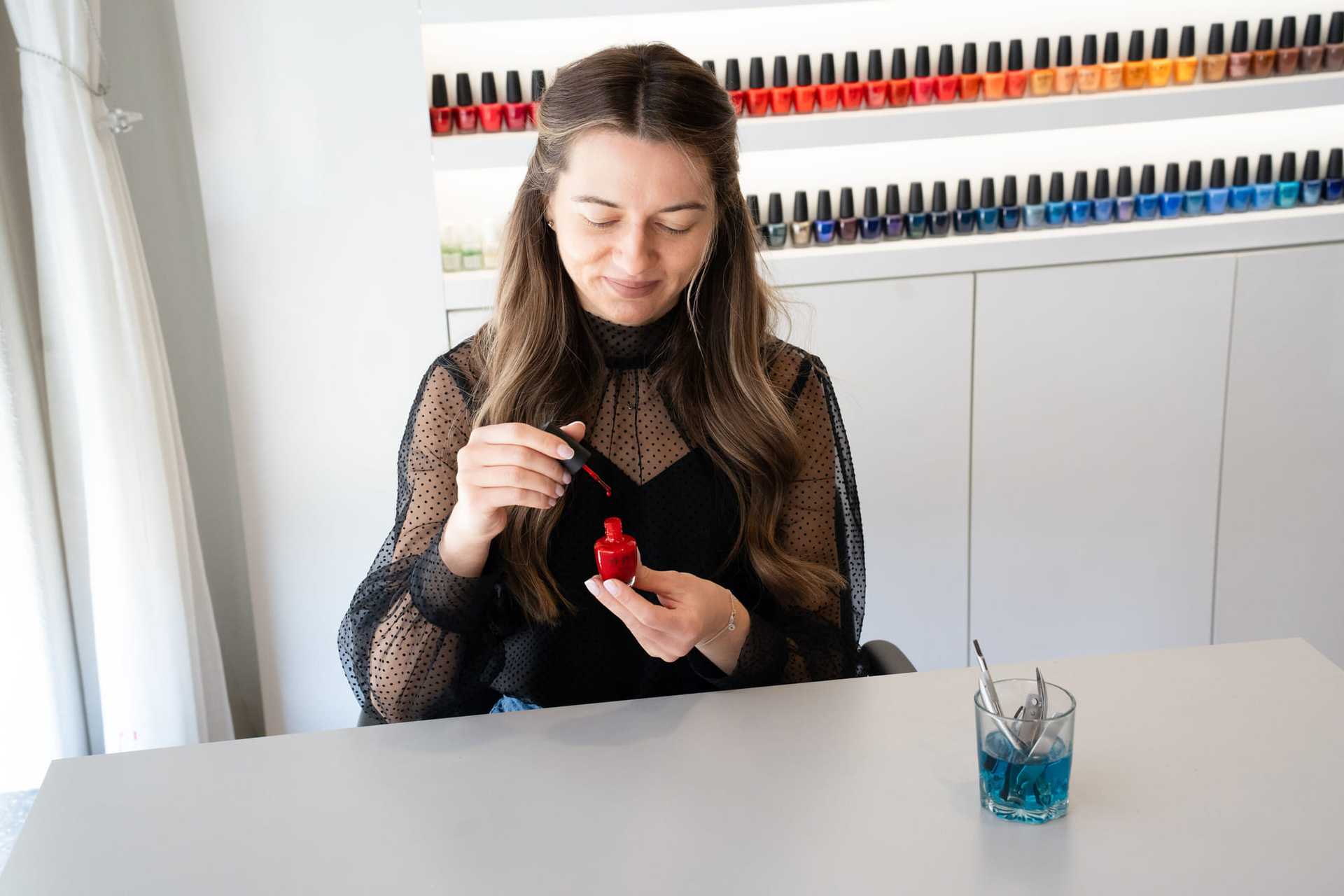Woman painting nails with red polish at a salon, colorful bottles on shelf behind.