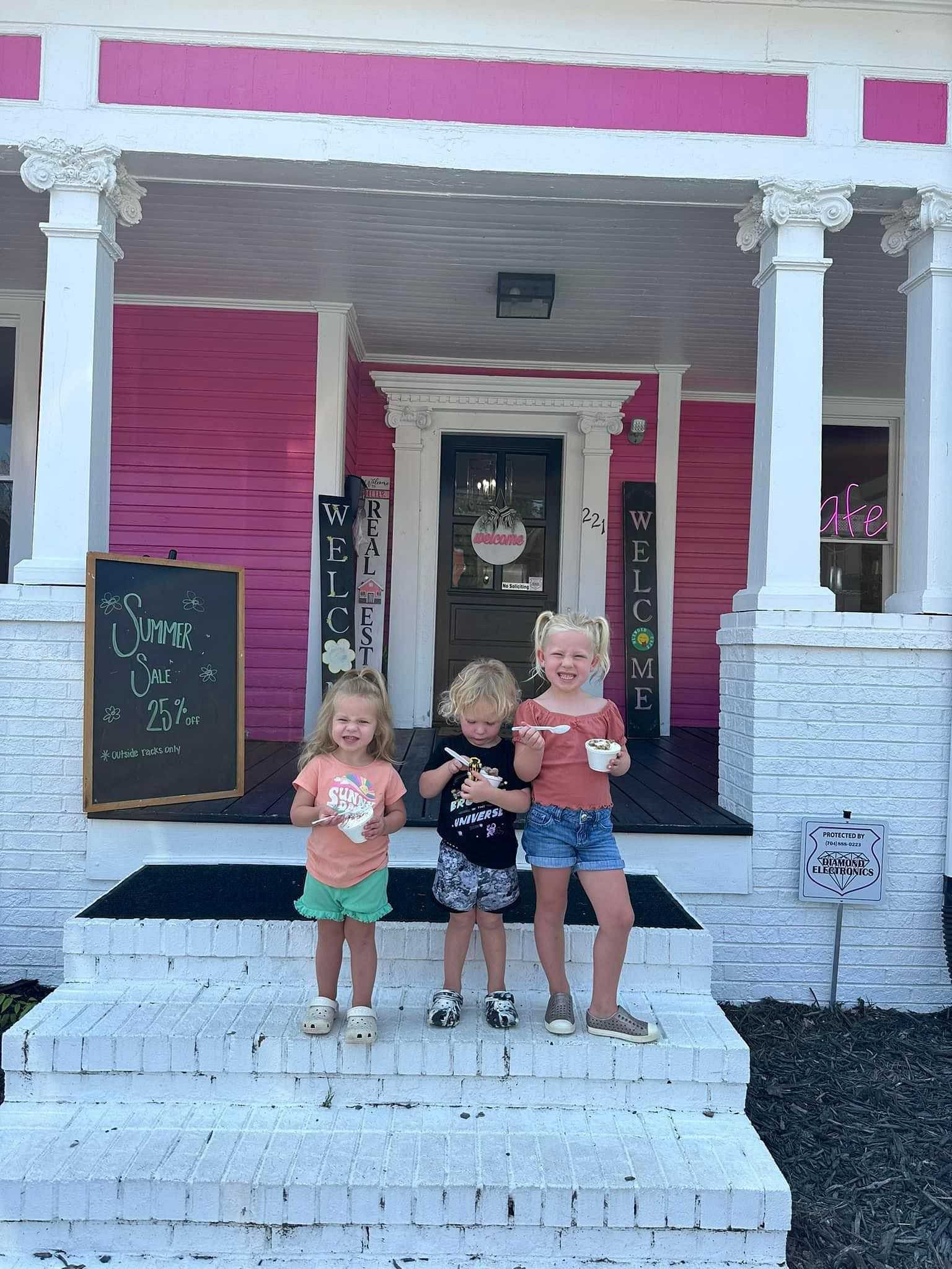 Three children enjoying ice cream on the front steps of a pink and white building with sale signs.