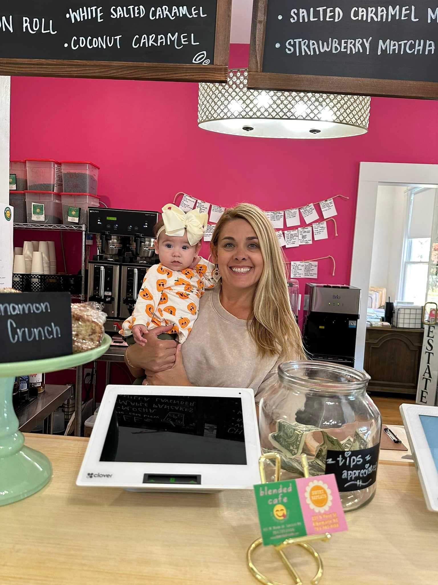 Woman holding a baby in a Halloween outfit behind a coffee shop counter with menu boards above.