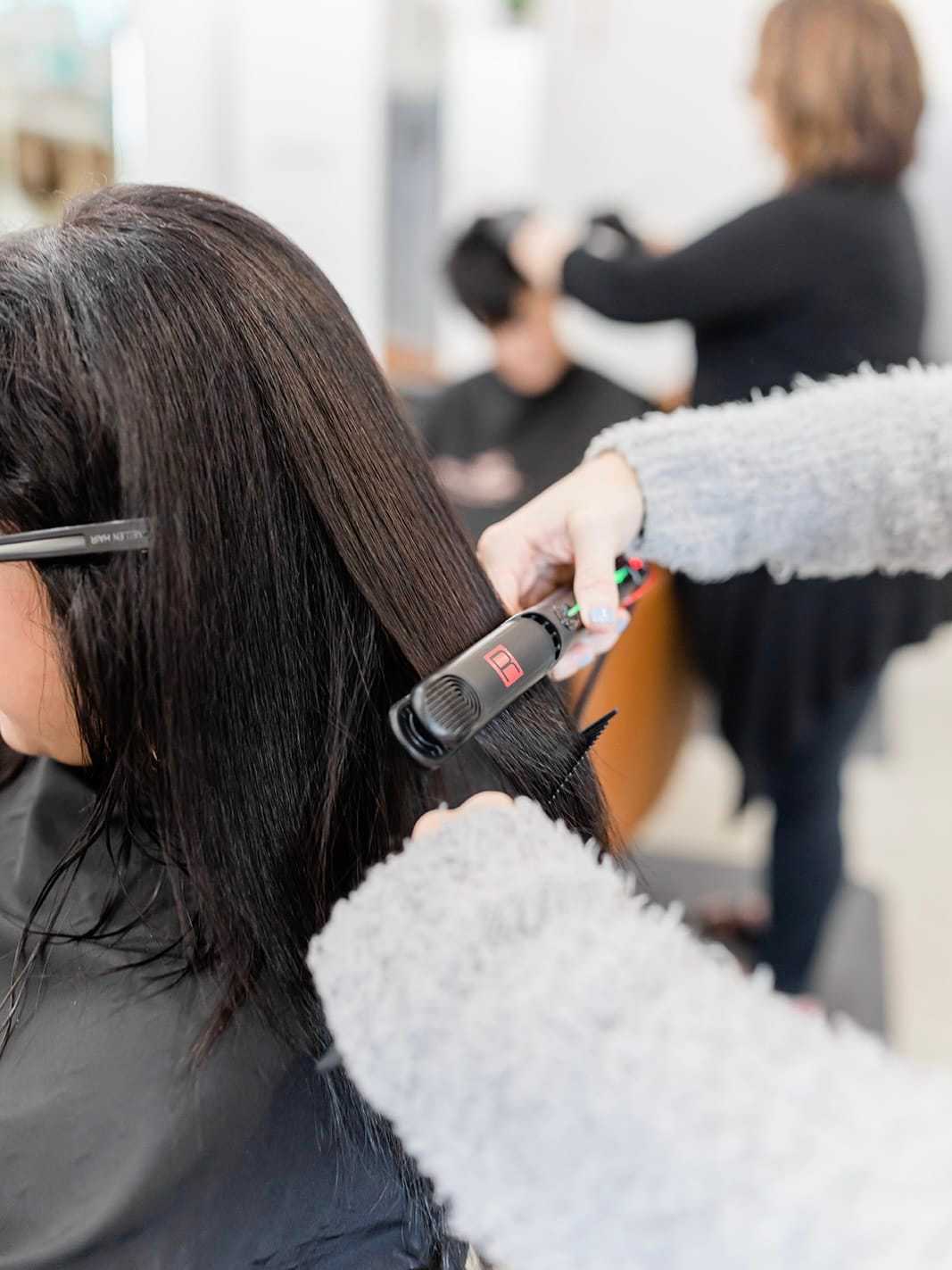Hairdresser straightening a client's long dark hair in a salon setting.