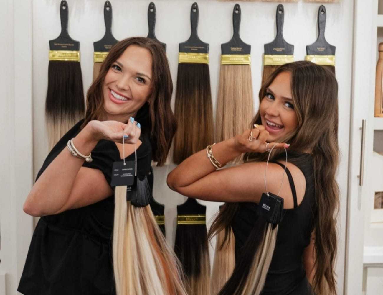 Two smiling women holding hair extensions in a salon display area.