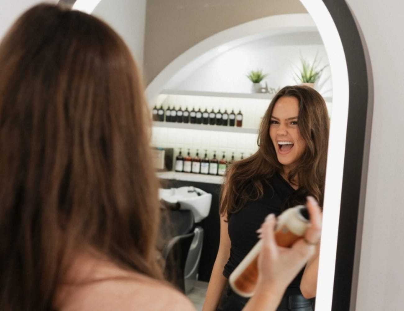 Woman smiling in mirror, holding hairspray in a stylish salon with product shelves.