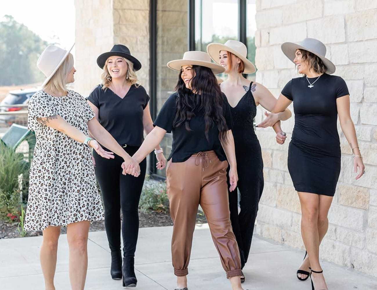 Five women walking together, smiling, each wearing stylish hats and casual attire.