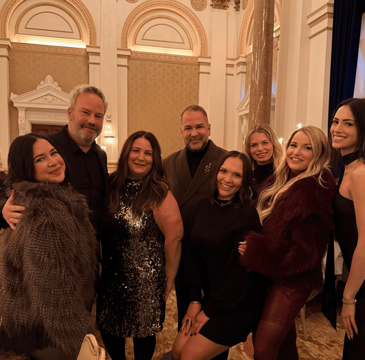 Group of people smiling at a formal event indoors with elegant decor in the background.