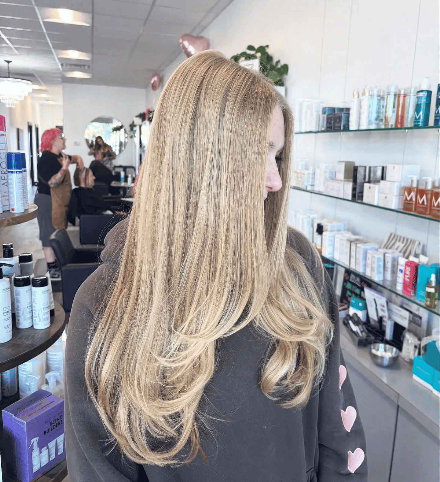 Blonde woman with long hair in a salon, wearing a hoodie with heart patches.