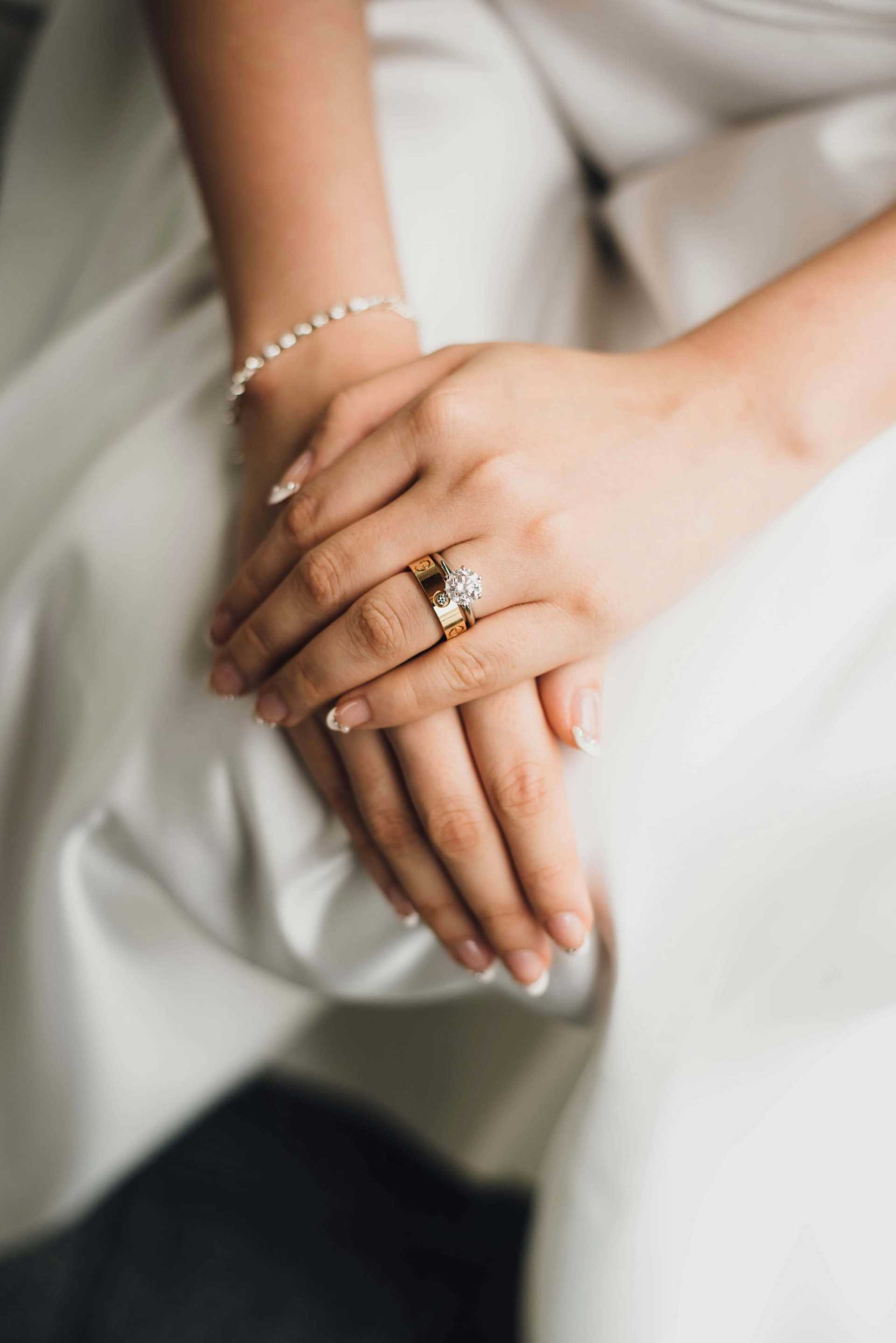 Bride's hands with wedding and engagement rings on a white dress.