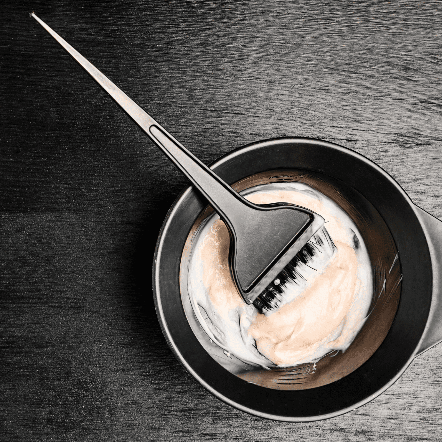 Hair dye brush in bowl with cream mixture on dark wooden surface.