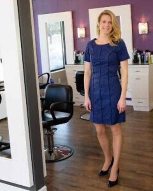 A woman standing in a salon, wearing a blue dress and black heels, with styling chairs nearby.