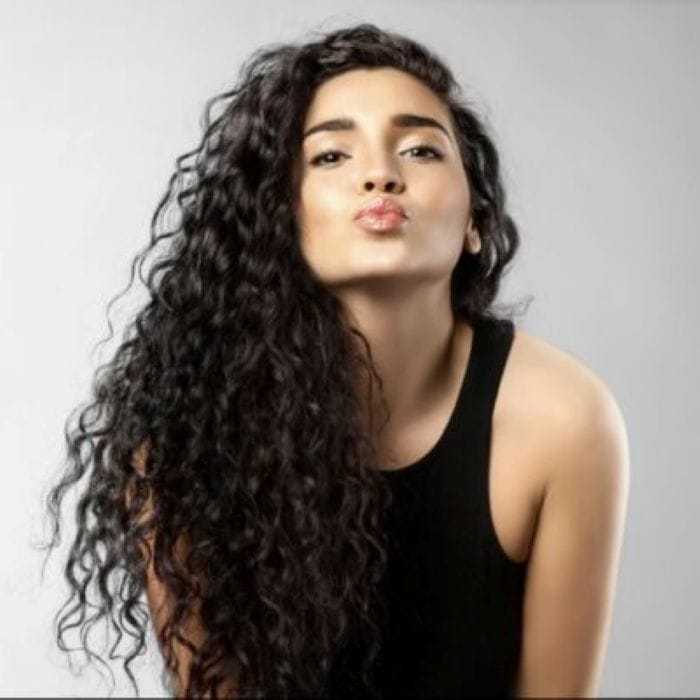 A person with long curly hair poses against a plain background, blowing a kiss.