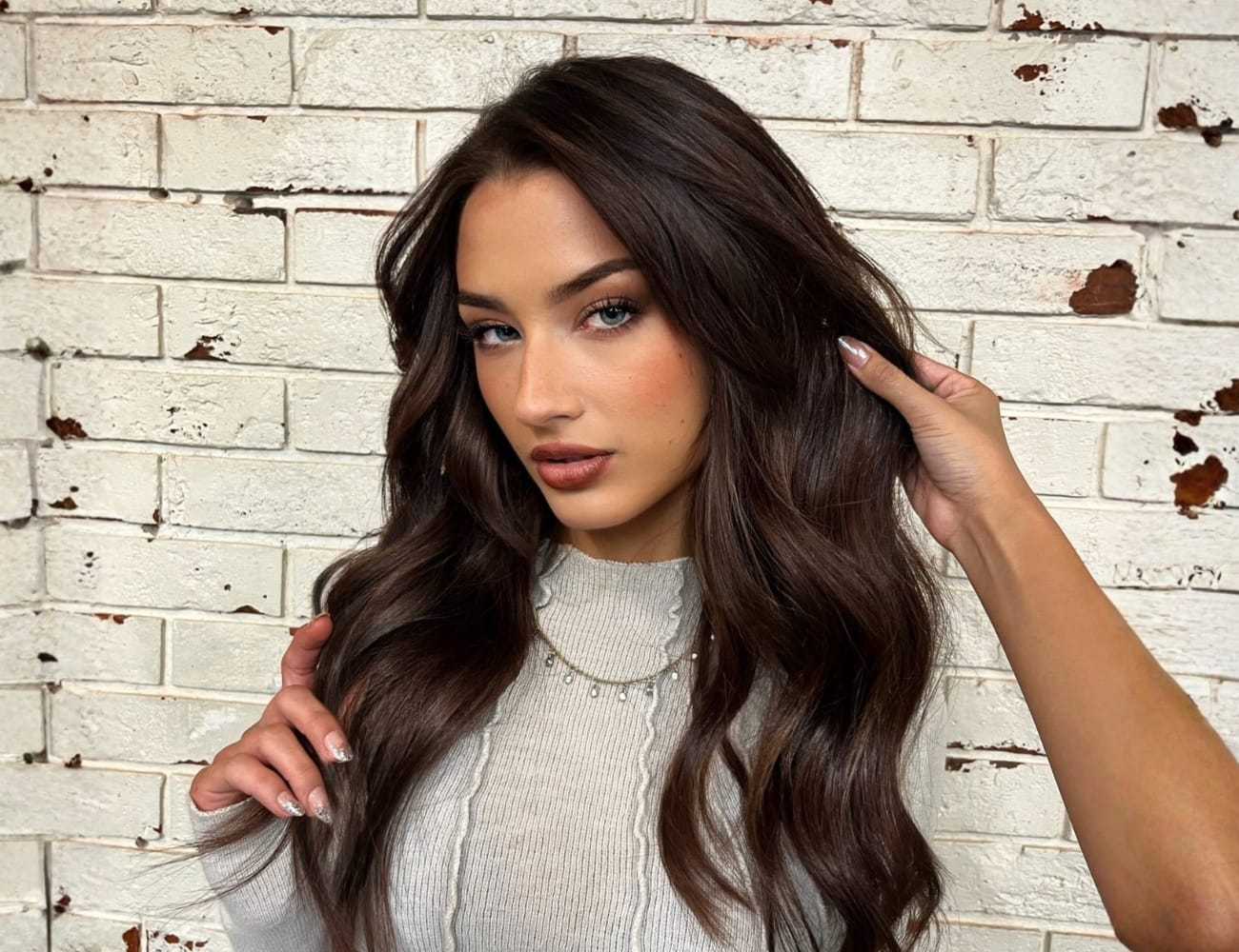 Woman with long brown hair posing against a white brick wall.