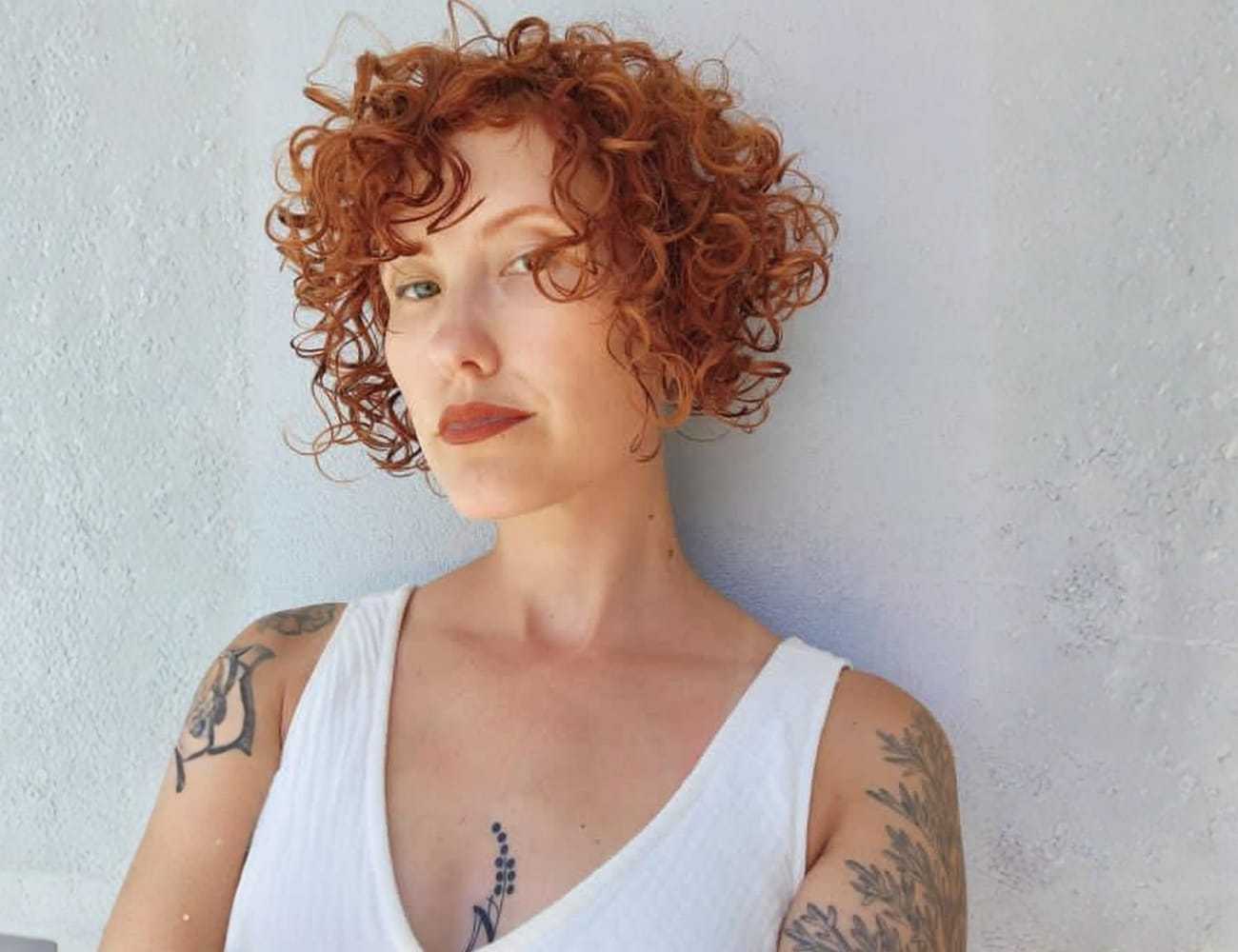 Red-haired woman with tattoos poses against a light background, wearing a white top.