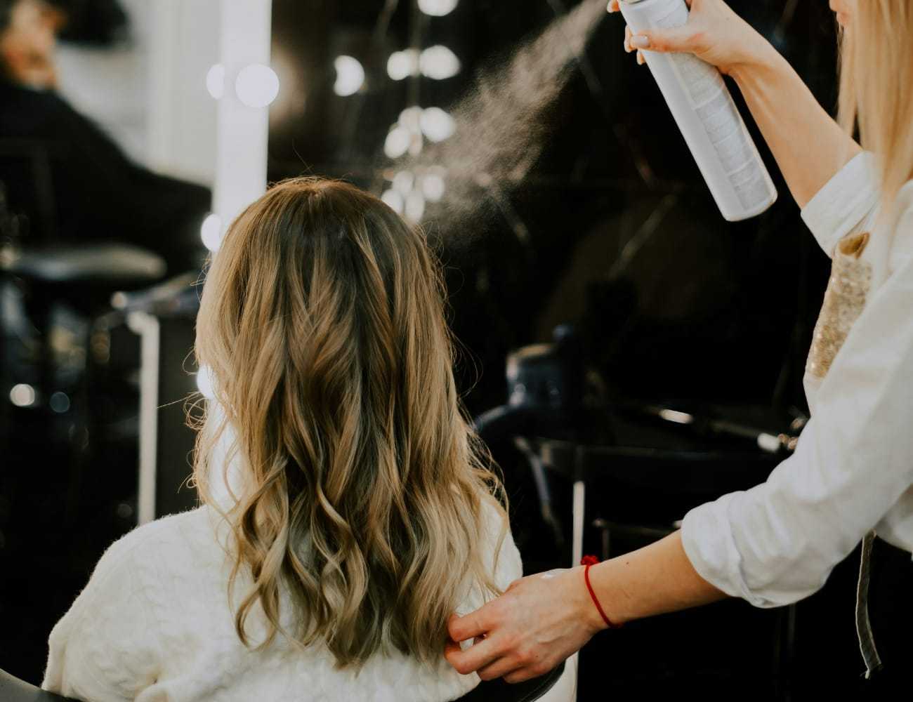 Hairdresser styling woman's hair with spray at salon.