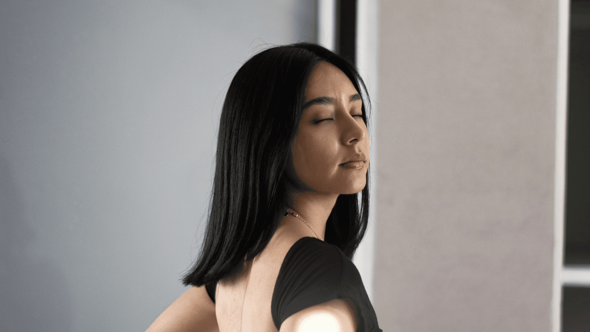 Woman with closed eyes and long hair, standing against a neutral background.