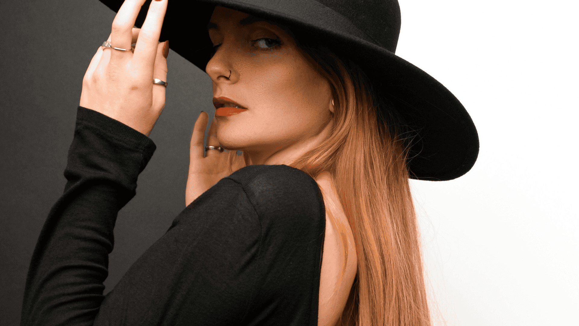 Woman in a black hat and dress, looking sideways with red lipstick and long hair.