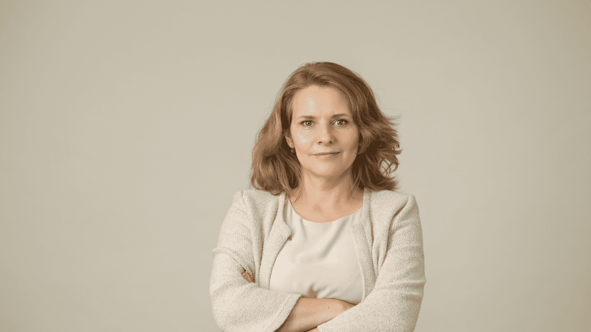 Woman with crossed arms in a light jacket, smiling confidently against a neutral background.