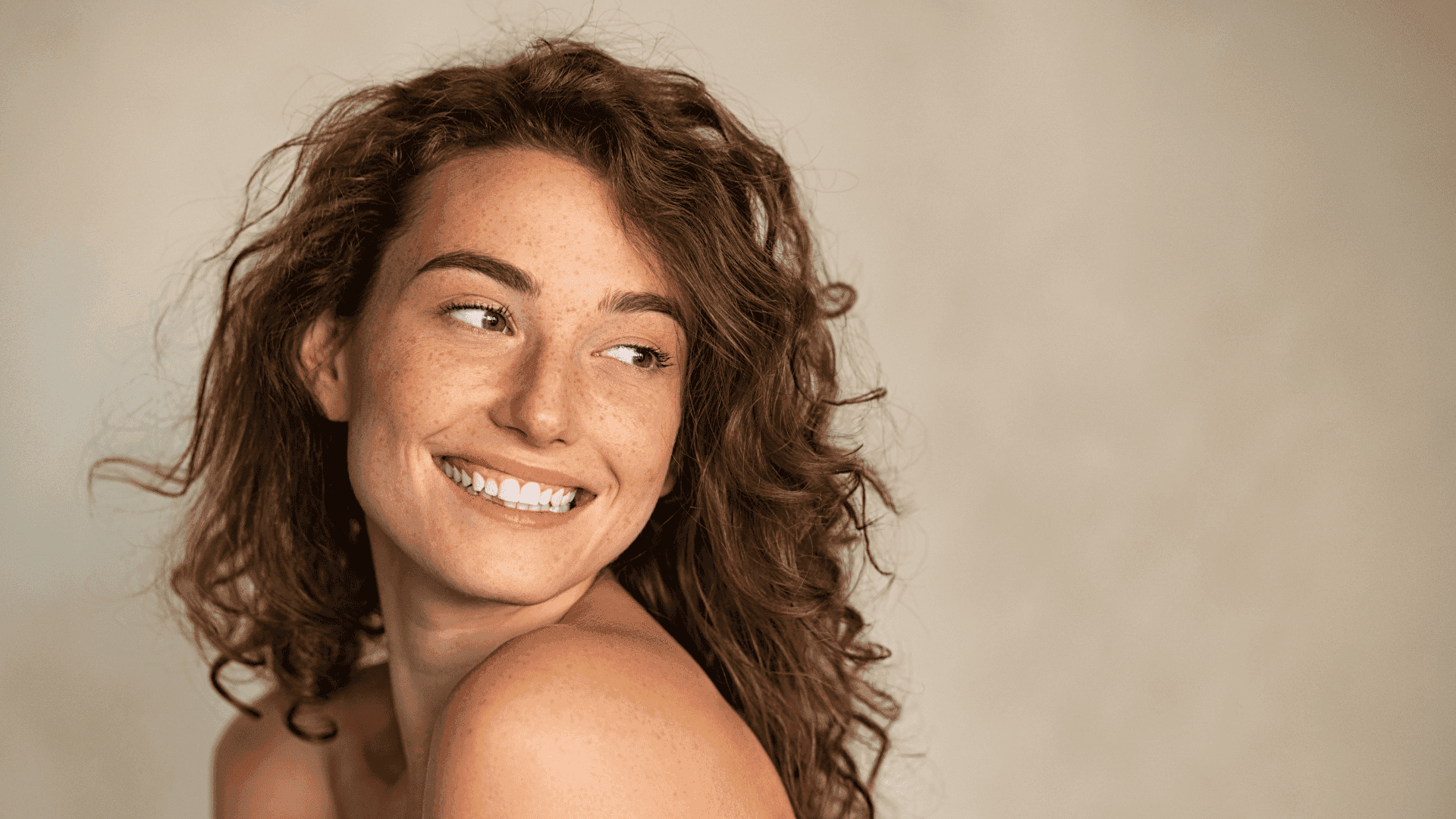 Smiling woman with long curly hair looking to the side against a neutral background.