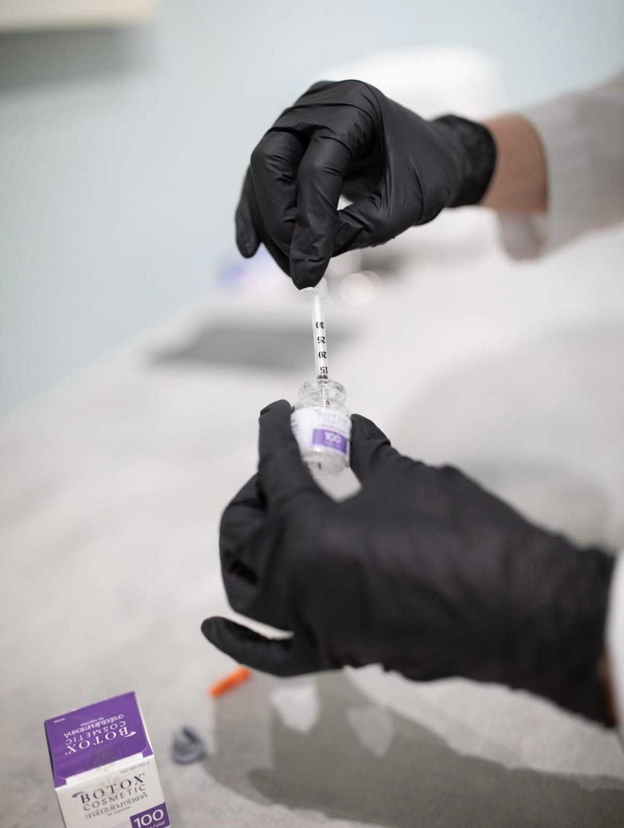 Hands in black gloves preparing a syringe with a Botox vial on a medical surface.