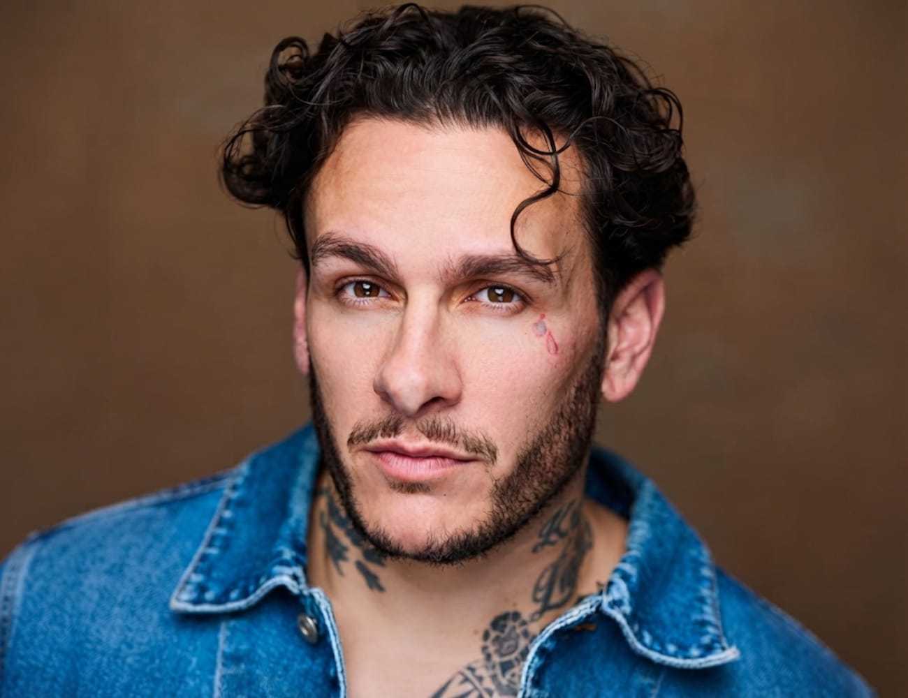 Man with tattoos and curly hair wearing a denim shirt, looking directly at the camera.