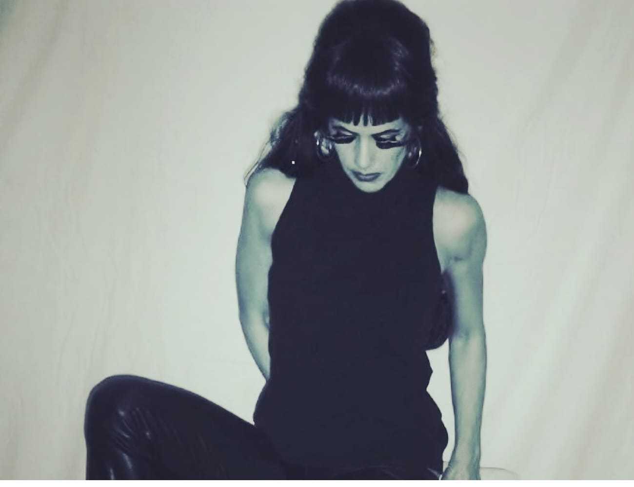 Female wearing a sleeveless top, seated against a plain backdrop, eyes downcast.