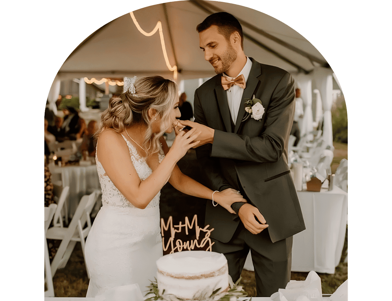 Newlyweds share cake at outdoor reception under a tent.
