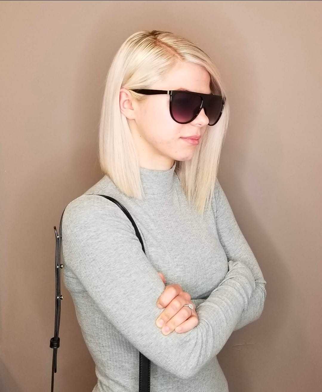 Short-haired person in sunglasses, wearing a gray top, standing with arms crossed against a beige wall.