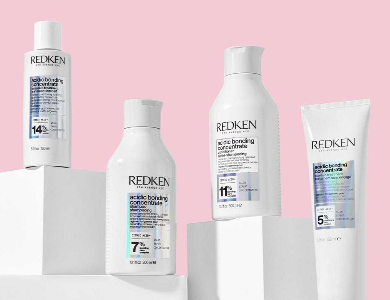 Four Redken hair care bottles on white platforms against a pink background.