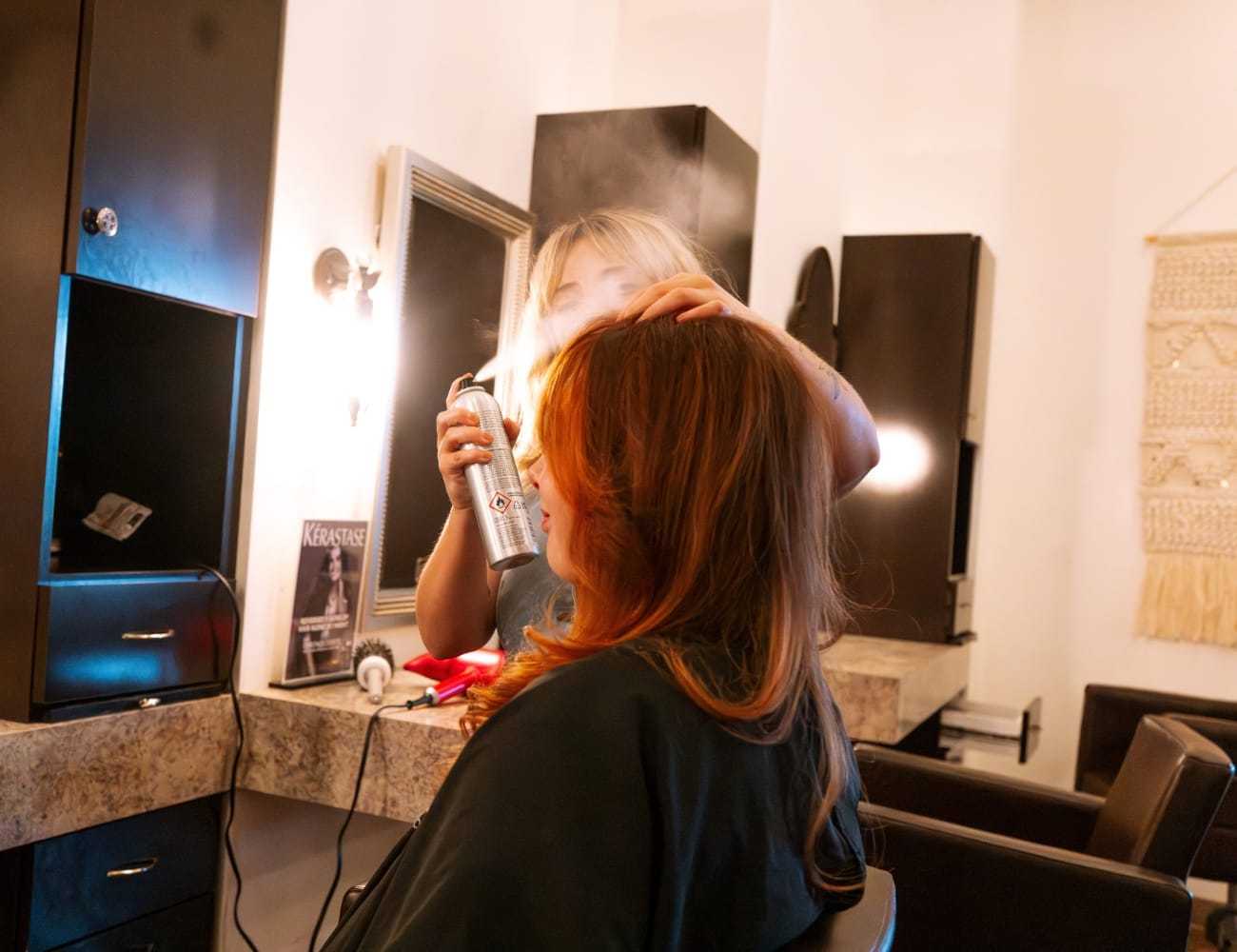 Hairdresser styling a client's red hair with hairspray in a salon.