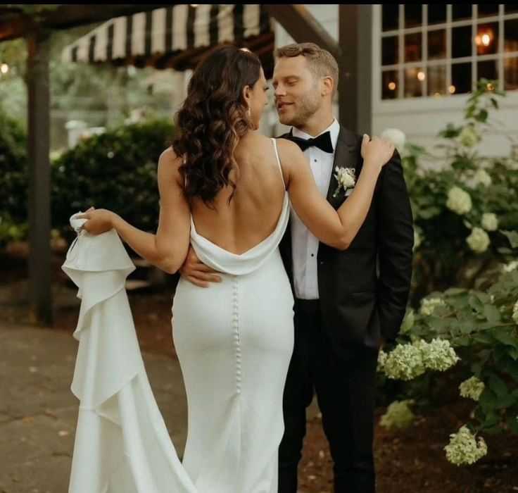 Bride and groom embrace outdoors, bride in backless gown, groom in tuxedo.