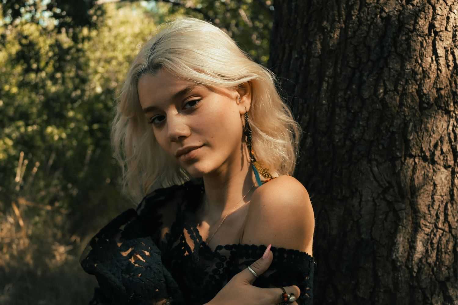 Young woman with blonde hair, wearing a black lace dress, standing by a tree in a forest.