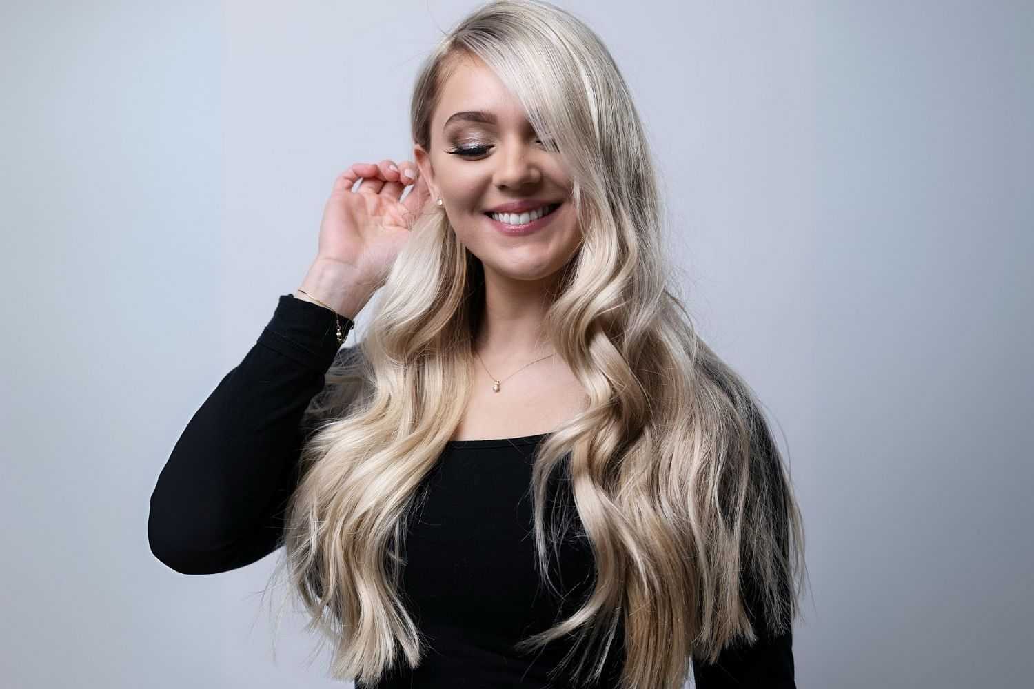Smiling woman with long, wavy blonde hair, wearing a black top against a simple background.