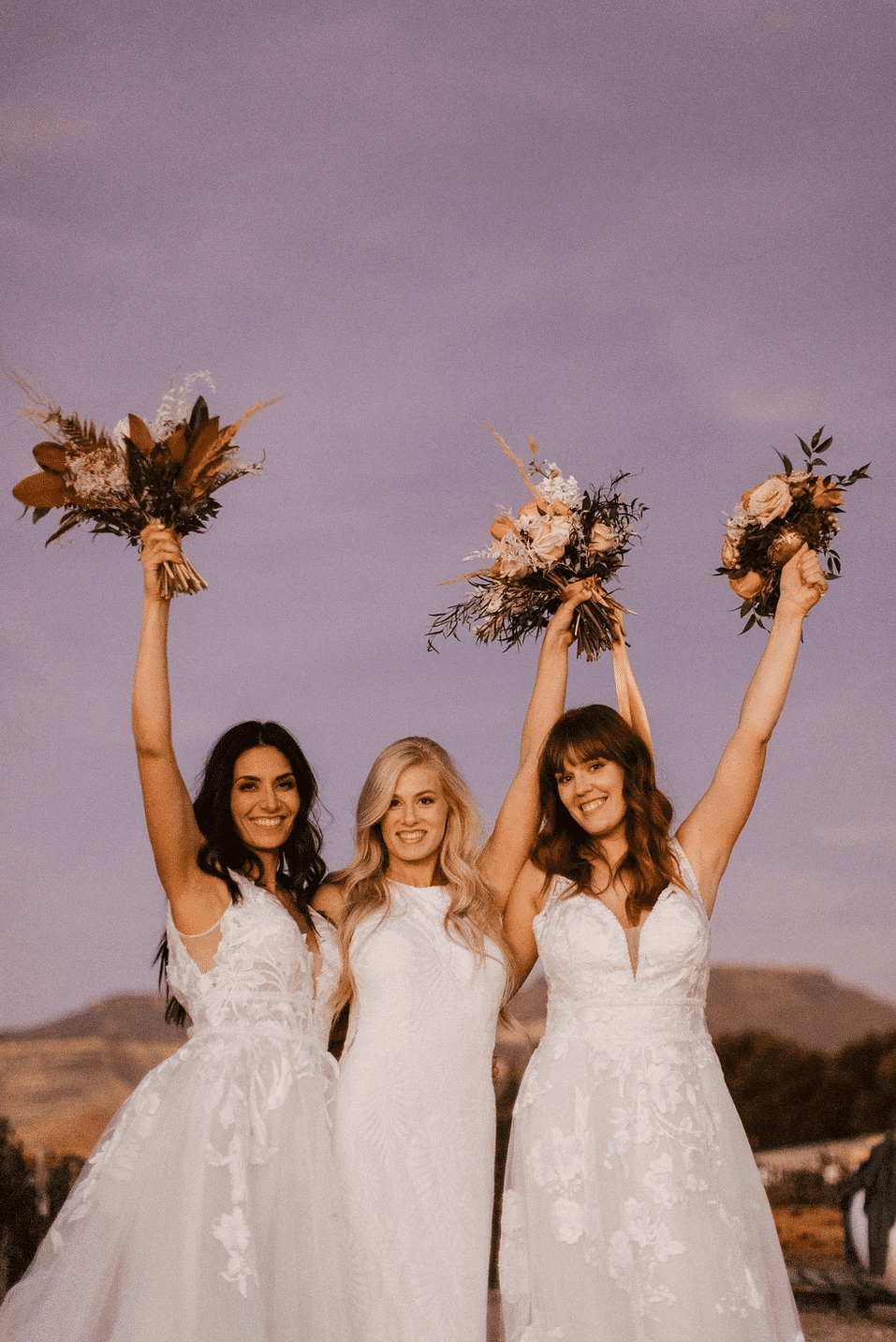 Three brides in white dresses holding flower bouquets, smiling and celebrating against a scenic backdrop.