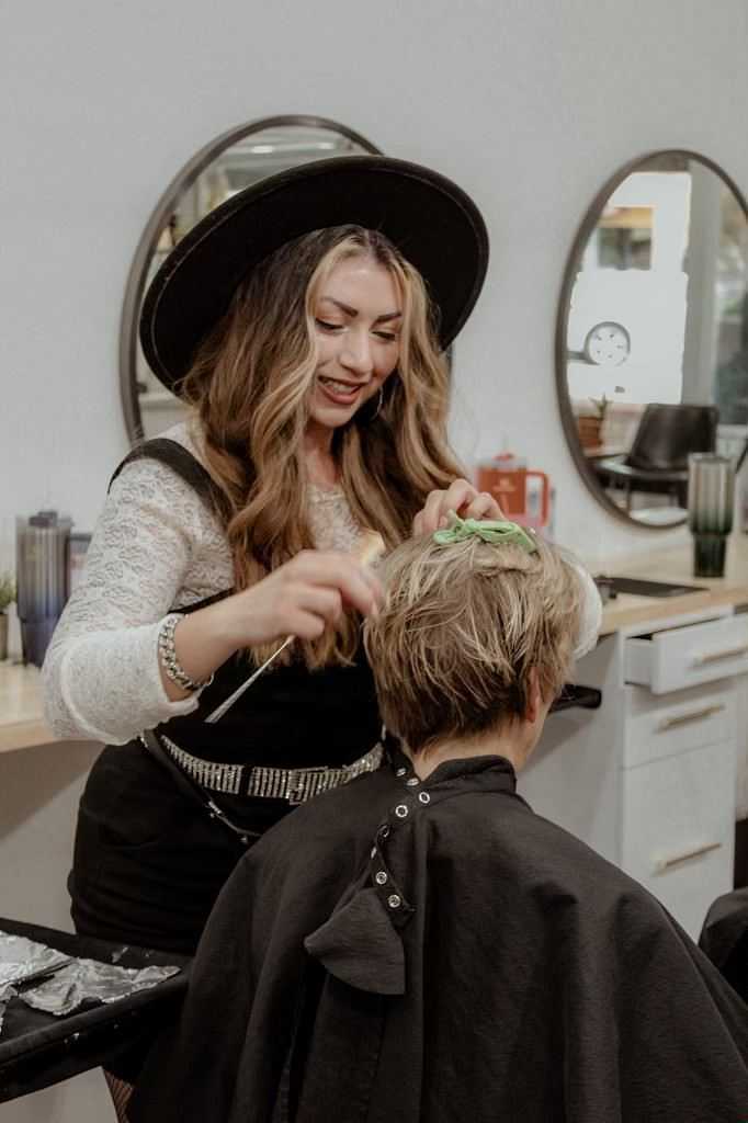 Hairdresser styling a client's short hair in a salon with round mirrors and hair products in the background.