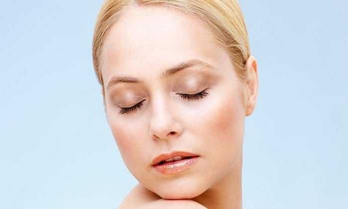 Woman with closed eyes and soft makeup, resting her chin on her hand against a light blue background.