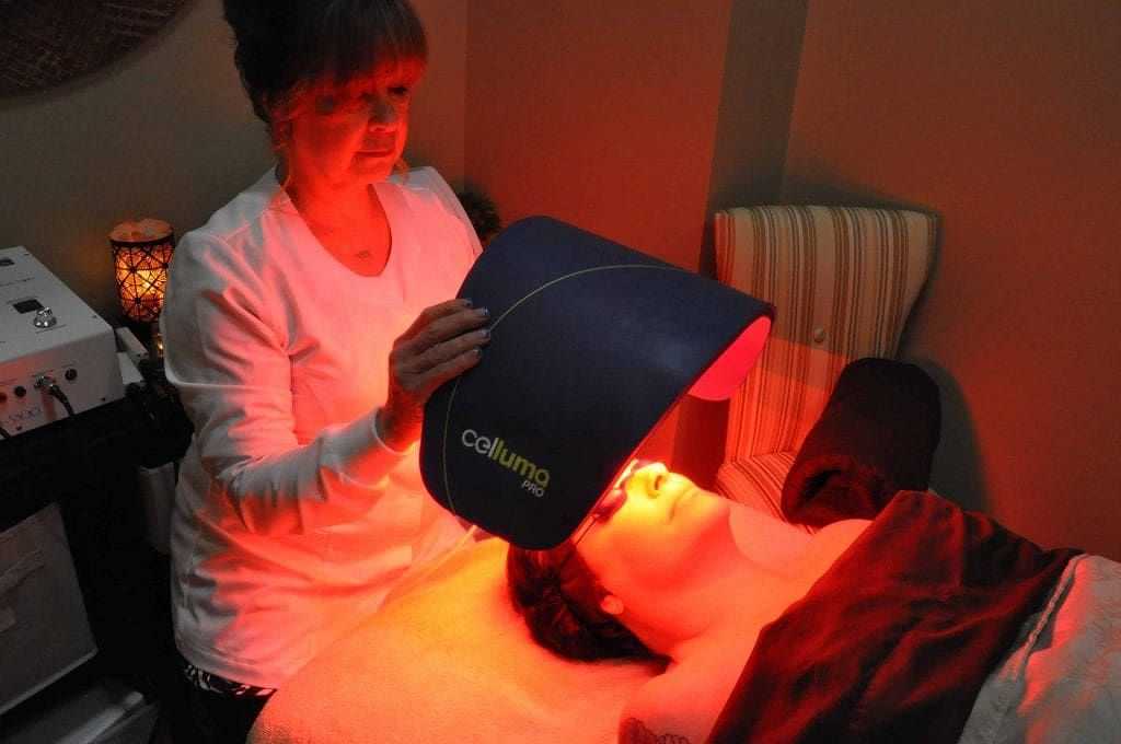 A person receiving a facial light therapy treatment from a practitioner in a dimly lit room.