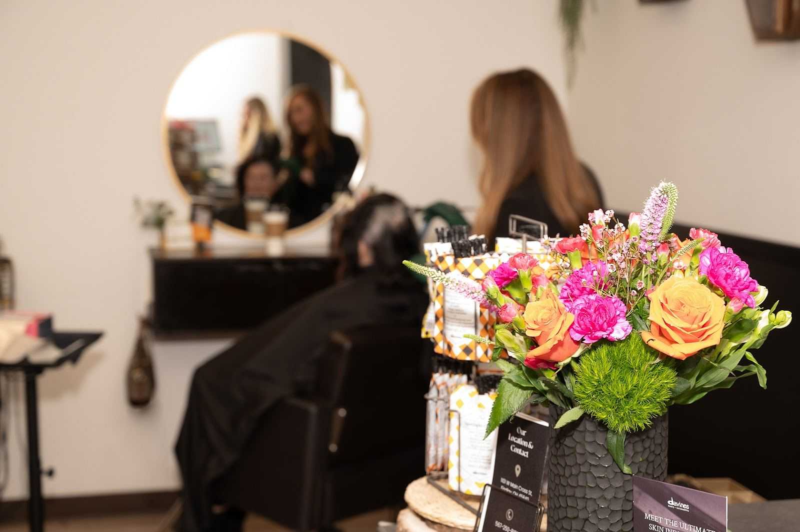 Hair salon with flowers, a stylist, and a seated client reflected in a round mirror.