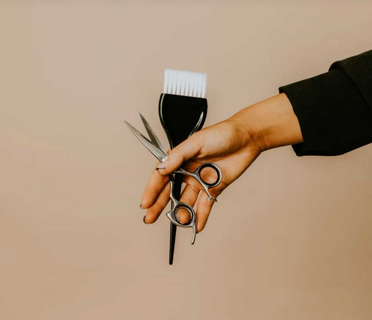 Hand holding scissors and hair dye brush against a beige background.