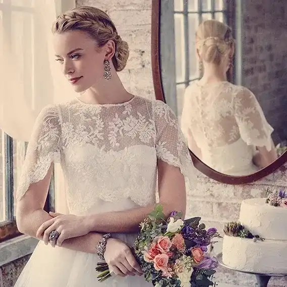 Bride in lace gown holding bouquet, standing by window with reflection in mirror.