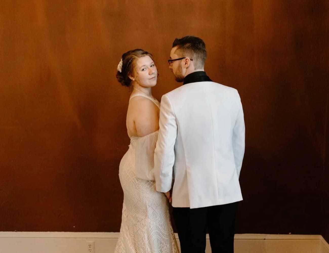 Bride and groom in elegant attire, posing against a warm brown background.