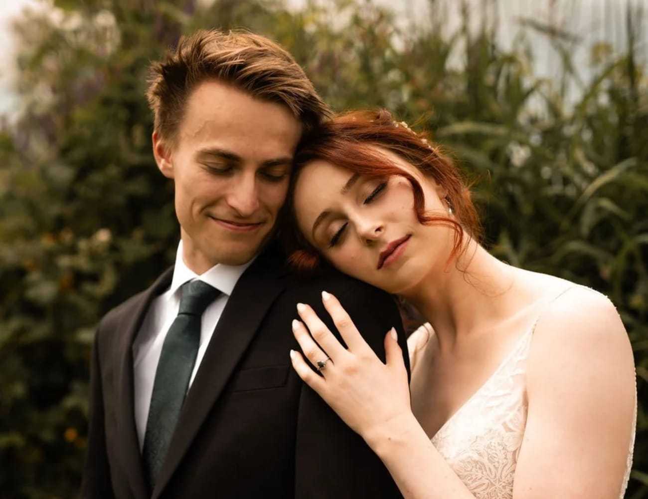 Couple embraces in garden, woman shows engagement ring while leaning on man's shoulder.