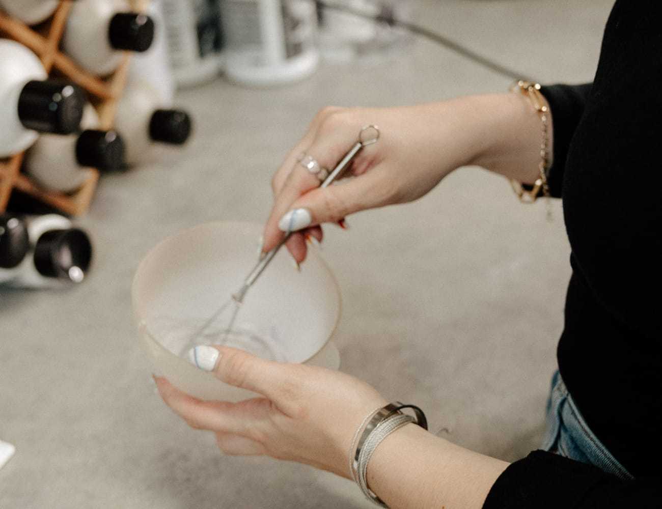 Woman mixing hair dye in a bowl with a whisk, bottles in the background.