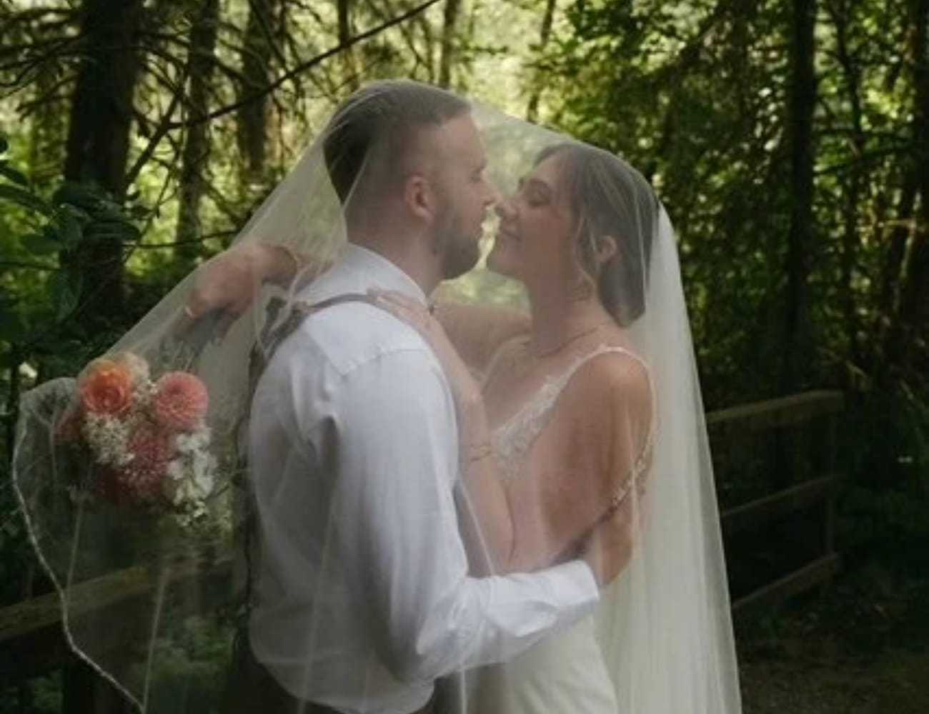Bride and groom kiss under a veil in a forest setting, holding flowers.