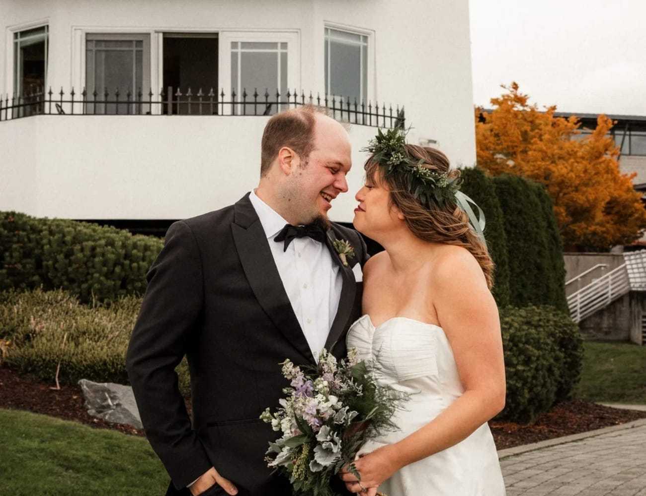 Bride and groom smiling at each other in front of a white building and autumn trees.