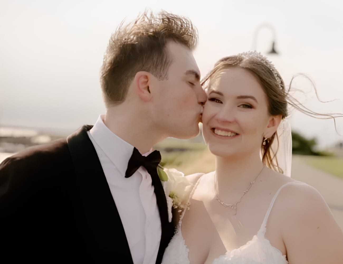 Couple in wedding attire smiling as the groom kisses the bride on the cheek.