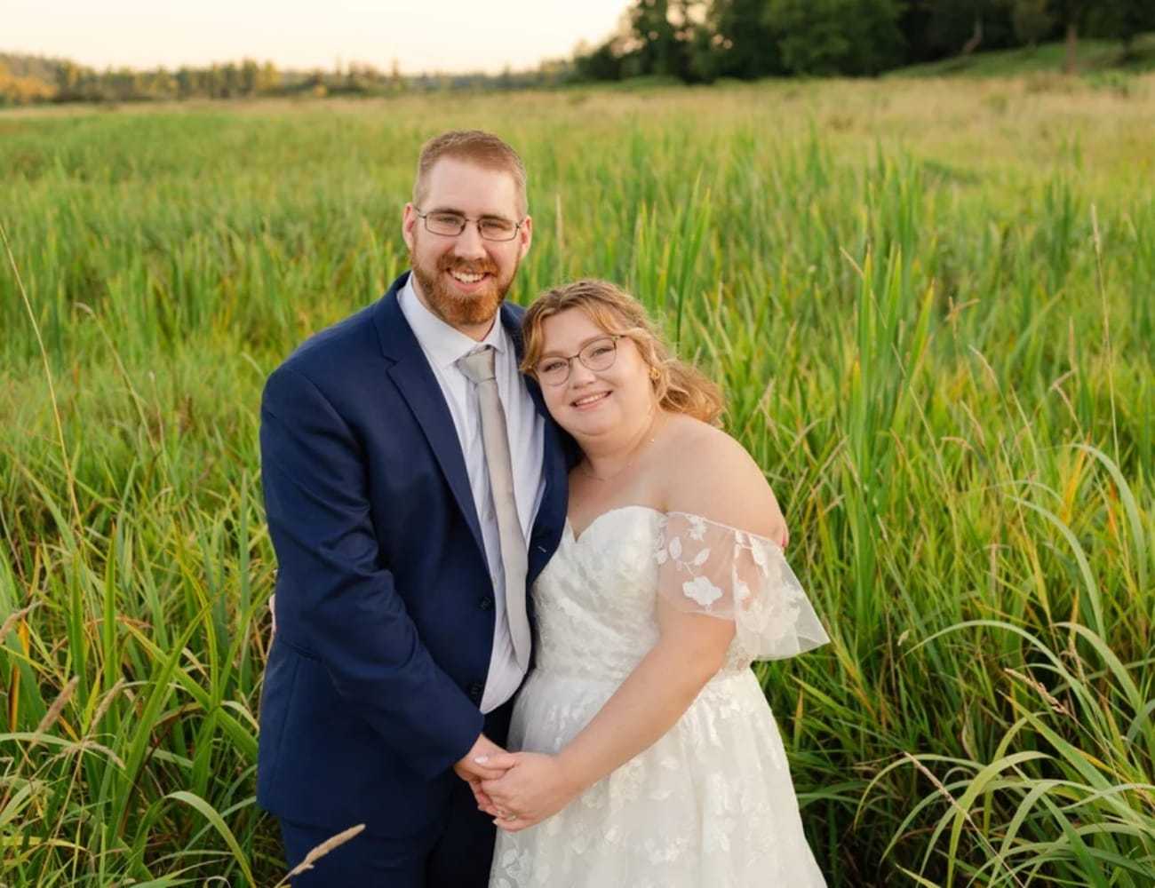 A couple in wedding attire smiling in a lush, green field.