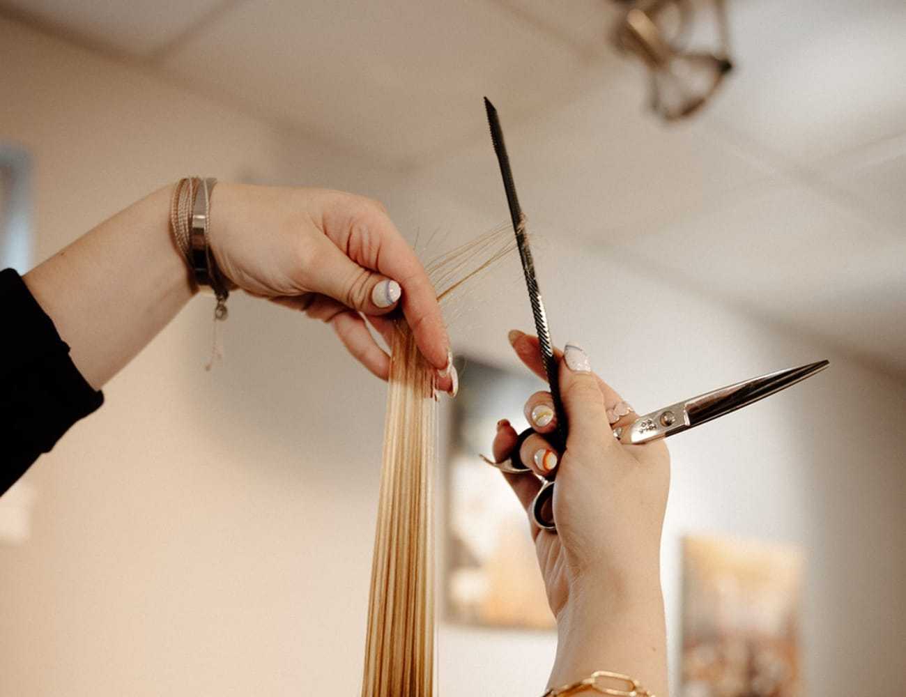 Hairdresser cutting client's long blond hair with scissors and comb.