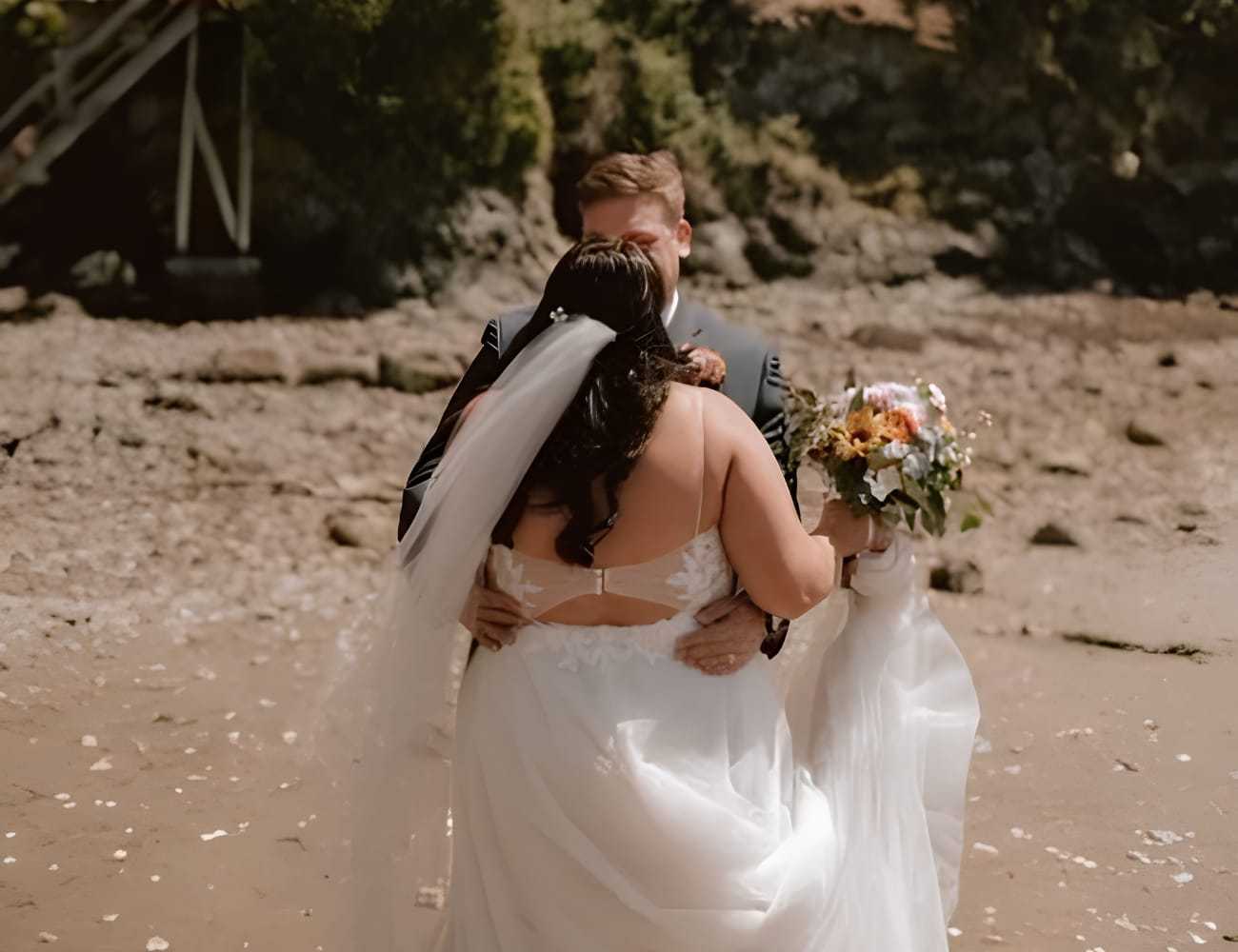 Bride and groom embrace on a rocky beach, holding a colorful bouquet.