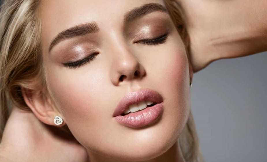 Close-up of a woman with closed eyes and neutral makeup, hands touching her head.