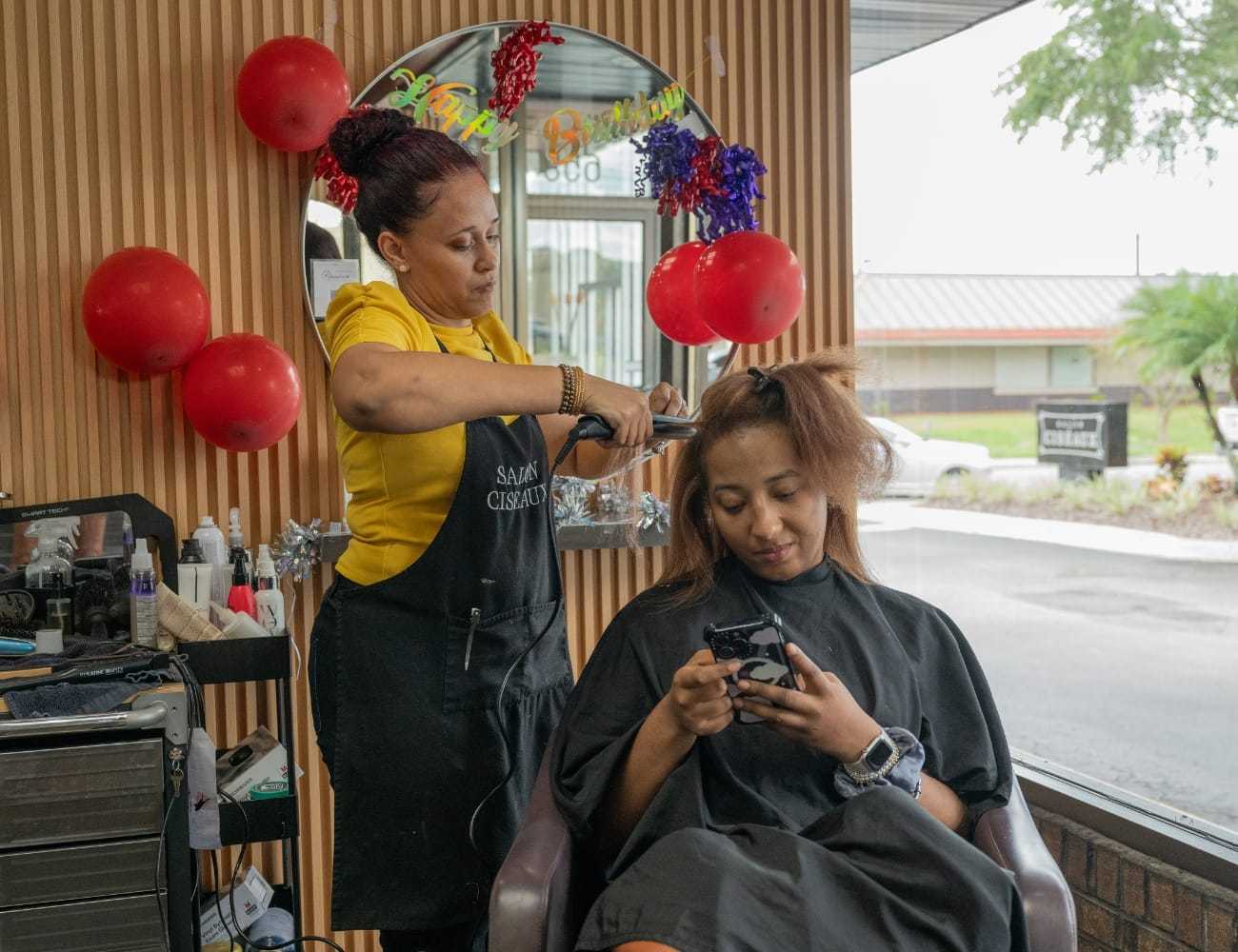 Hairdresser styling customer's hair while client uses phone in a salon, with balloons in view.