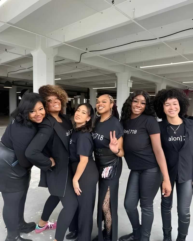 Group of six smiling people in matching black Salon 718 shirts in an industrial space.
